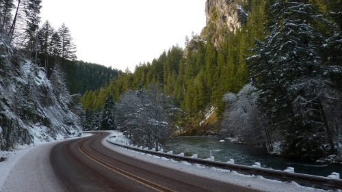 Winding road through a snowy landscape with tall trees and a rocky mountain in the background.