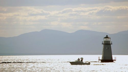 A small boat sails near a lighthouse on a shimmering lake, with mountains and cloudy skies in the background.