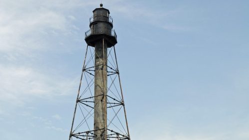 A tall, black lighthouse tower stands beside a small, weathered building in a grassy field under a blue sky.