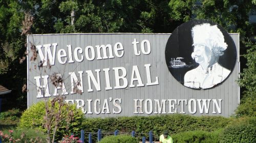 Sign welcoming visitors to Hannibal, featuring a portrait and the phrase "America's Hometown" amidst greenery.