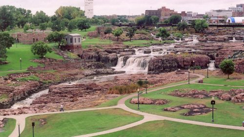 A scenic view of a park featuring waterfalls, rocky terrain, and green spaces under a cloudy sky.