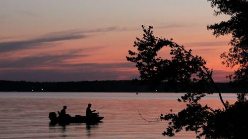 Silhouetted fishermen in a boat on a calm lake at sunset, with vibrant colors reflecting on the water.