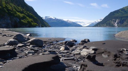 A serene landscape featuring a calm blue lake surrounded by mountains and a rocky shoreline under a clear sky.