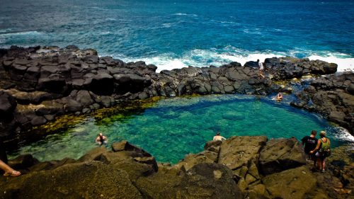 A natural pool surrounded by rocky shores, with people swimming and enjoying the ocean view under a clear blue sky.