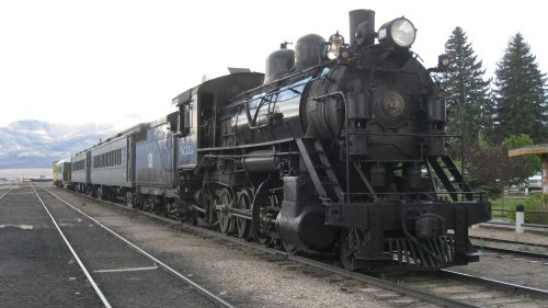 A vintage steam locomotive with a passenger car, parked on tracks with trees and mountains in the background.