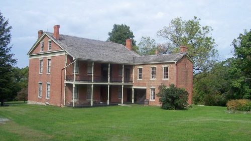 Historic brick house with a porch, surrounded by green grass and trees under a cloudy sky.