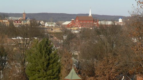 A scenic view of a small town with church steeples, trees, and distant hills under a clear sky.