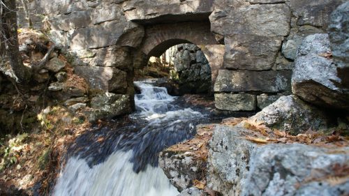 A stone arch bridge over a flowing stream, surrounded by rocks and autumn leaves.