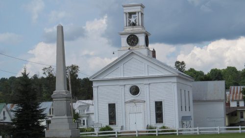 A white church with a clock tower and an obelisk monument in front, set against a cloudy sky and green landscape.