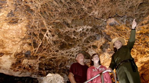 A park ranger points out features on a cave ceiling to two visitors, surrounded by intricate rock formations.