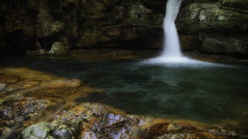 A serene waterfall cascades into a clear pool, surrounded by rocky terrain and lush greenery.