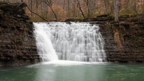 A serene waterfall cascades over rocky ledges into a tranquil, green pool surrounded by trees.