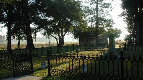 A serene cemetery with a wrought iron fence, surrounded by trees and a misty landscape in the background.