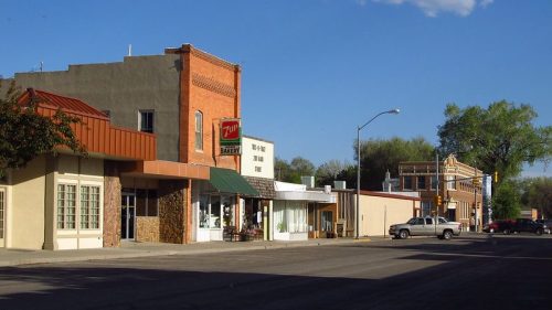 A quiet street scene featuring brick buildings, shops, and a clear blue sky with a few clouds.