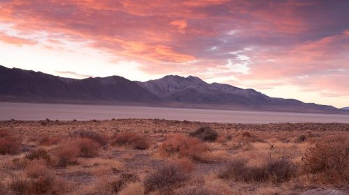 A desert landscape at sunset, featuring mountains in the background and colorful clouds in the sky.