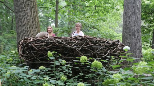 Two women stand inside a large, woven nest structure surrounded by lush greenery and trees.