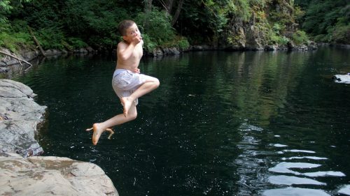 A boy jumps joyfully into a calm, green-tinted swimming hole surrounded by trees and rocky edges.