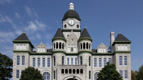 Historic stone building with a clock tower, featuring ornate architecture and green accents against a blue sky.