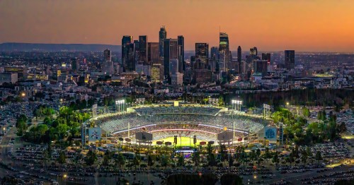 LA Dodger stadium at sunset with cars parks around, and the view of downtown LA in the background.