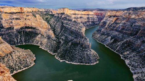 Aerial view of a winding river surrounded by steep, rocky cliffs and lush greenery under a cloudy sky.