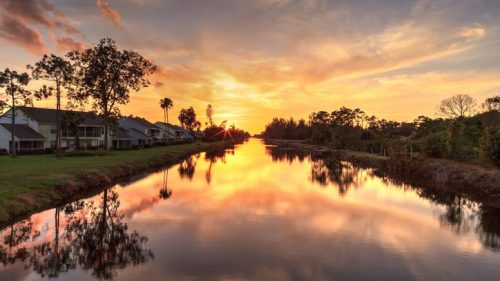 Sunset over a calm canal, reflecting vibrant colors and silhouettes of trees and houses along the water's edge.