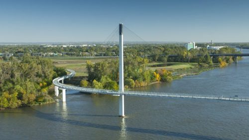 A modern cable-stayed bridge spans a river, surrounded by greenery and urban buildings in the background.
