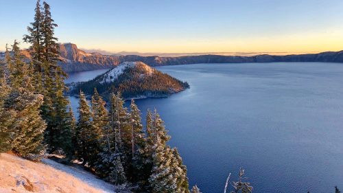 A serene view of Crater Lake surrounded by snow-capped trees and mountains under a clear blue sky at sunrise.