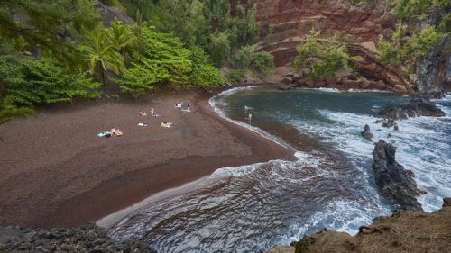 A scenic view of a red sand beach surrounded by lush greenery and rocky cliffs, with people relaxing by the water.