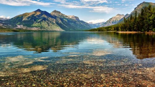 A serene lake surrounded by mountains, with clear water reflecting the sky and colorful pebbles visible beneath the surface.