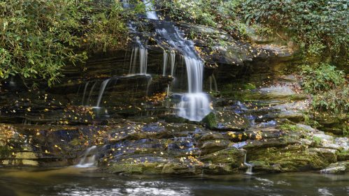 A serene waterfall cascades over rocky ledges, surrounded by lush greenery and autumn leaves.