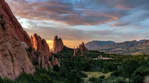 Sunset over rugged rock formations and lush greenery, with mountains in the background and a colorful sky.