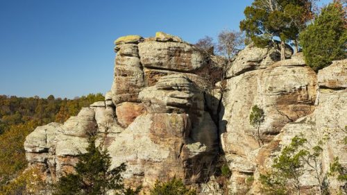Rocky cliffside with layered stone formations and sparse trees against a clear blue sky.