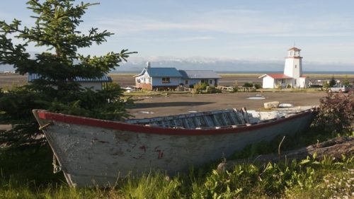 A weathered boat in the foreground with a lighthouse and buildings in the background, set against a scenic landscape.