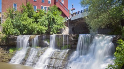 A serene waterfall cascades over rocks, surrounded by lush greenery and a red brick building in the background.