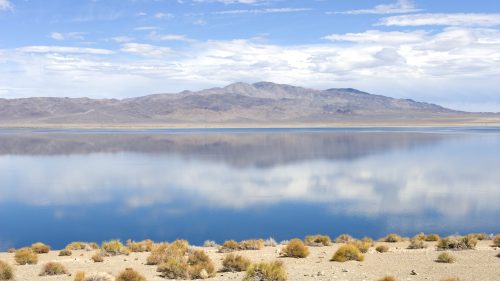 A serene landscape featuring a calm lake reflecting mountains and a blue sky, with sparse vegetation in the foreground.