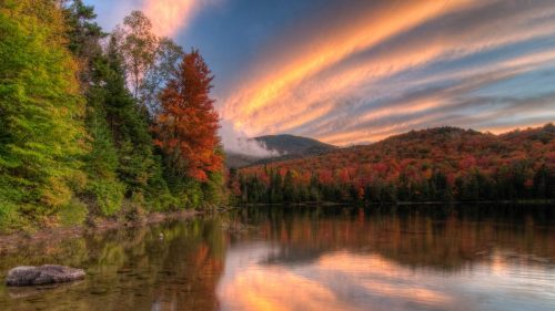 A serene lake surrounded by vibrant autumn foliage and a colorful sunset sky reflecting on the water's surface.