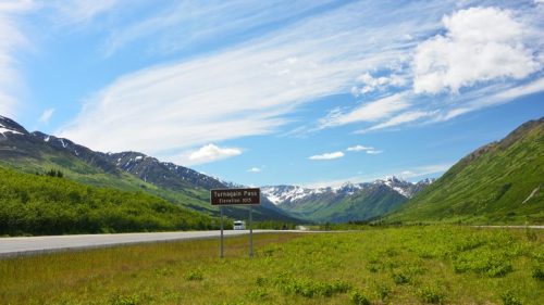 A scenic view of Turnagain Pass with mountains, greenery, and a blue sky dotted with clouds.