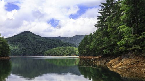 Serene lake surrounded by lush green mountains and trees under a partly cloudy sky. Reflections on the water's surface.