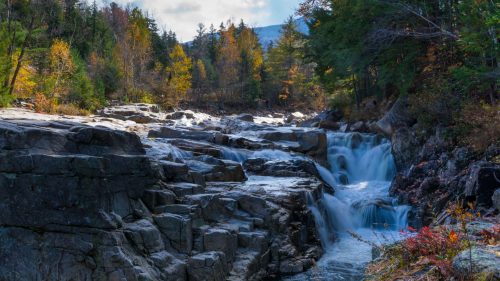 A serene waterfall cascades over rocky terrain, surrounded by autumn foliage and trees under a cloudy sky.