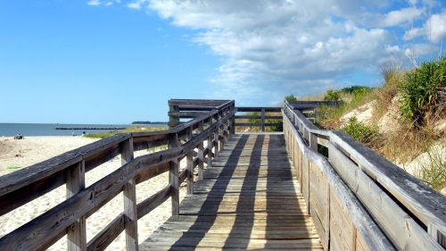 A wooden boardwalk leads to a sandy beach under a blue sky with scattered clouds.