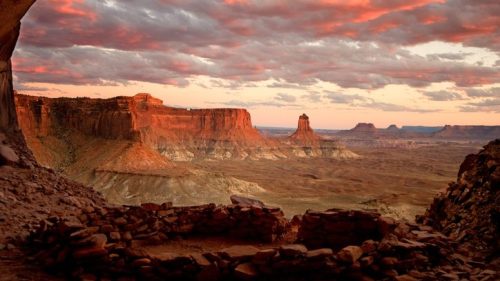 Sunset over a vast desert landscape with rock formations and dramatic clouds, viewed from a cave.