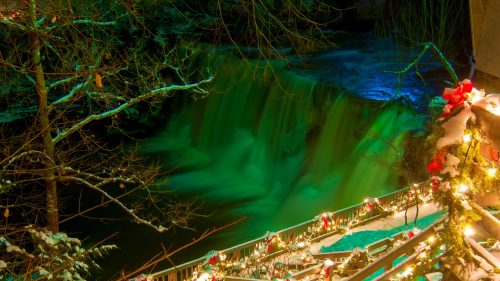 A snowy scene with a waterfall illuminated in green, surrounded by festive lights and decorations on a wooden railing.