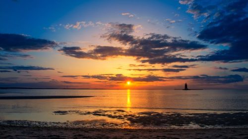 A serene sunset over calm waters, with clouds reflecting vibrant colors and a distant lighthouse silhouette.