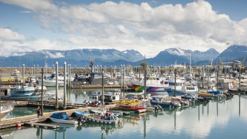 A bustling marina filled with various boats, surrounded by mountains and a partly cloudy sky.
