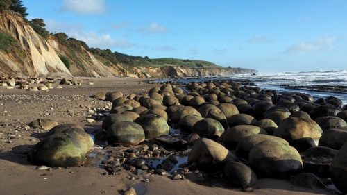 A rocky beach with large, round stones and cliffs in the background under a clear blue sky.