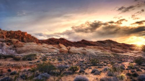 A scenic desert landscape at sunset, featuring rocky hills and sparse vegetation under a colorful sky.