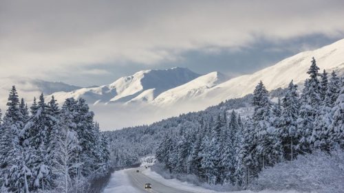 A snowy road winds through a forest, with majestic mountains in the background under a cloudy sky.