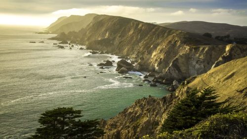 A scenic coastal view with rugged cliffs, gentle waves, and a golden sunset illuminating the landscape.