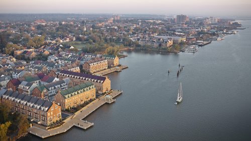 Aerial view of a waterfront town with colorful buildings, a marina, and a sailboat on the calm water.