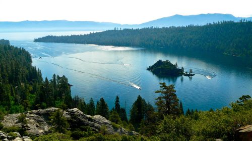 A serene lake surrounded by mountains and forests, with boats gliding across the water under a clear sky.